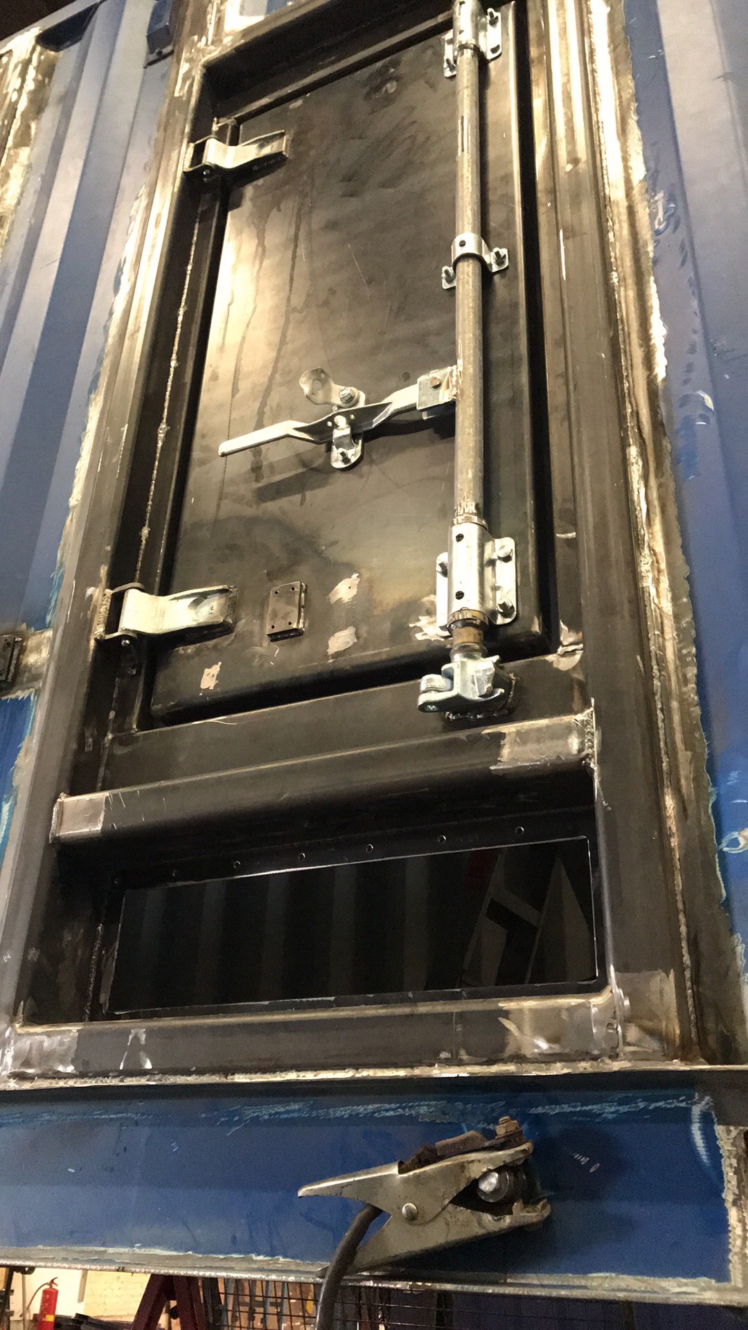 Metalowa rama okienna z zamkiem i zawiasami, w trakcie montażu w niebieskim kontenerze, widoczne ślady spawania i uchwyt spawalniczy.