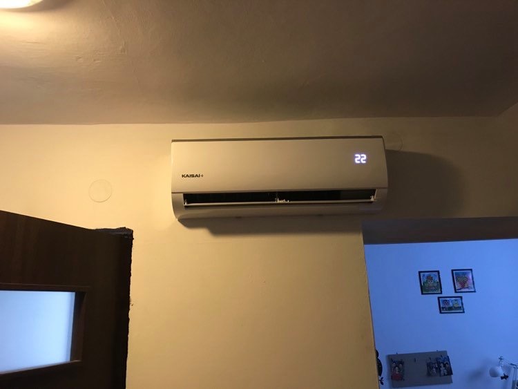 Biały klimatyzator Kaisai zamontowany na ścianie w pokoju, wyświetlający temperaturę 22 stopnie Celsjusza, widoczny fragment drzwi i ściana z dekoracjami w tle.