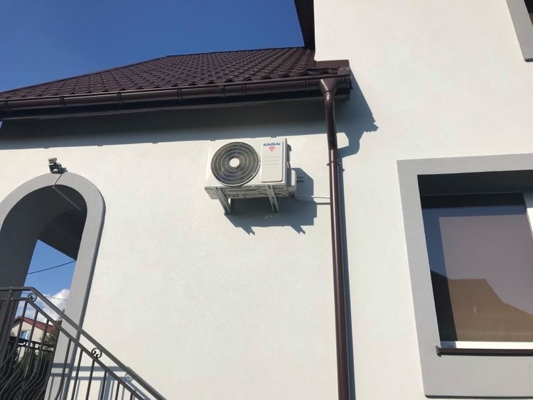 Biała jednostka zewnętrzna klimatyzacji marki Kaisai zamontowana na elewacji domu z brązowym orynnowaniem, widoczna z boku schodów wejściowych.