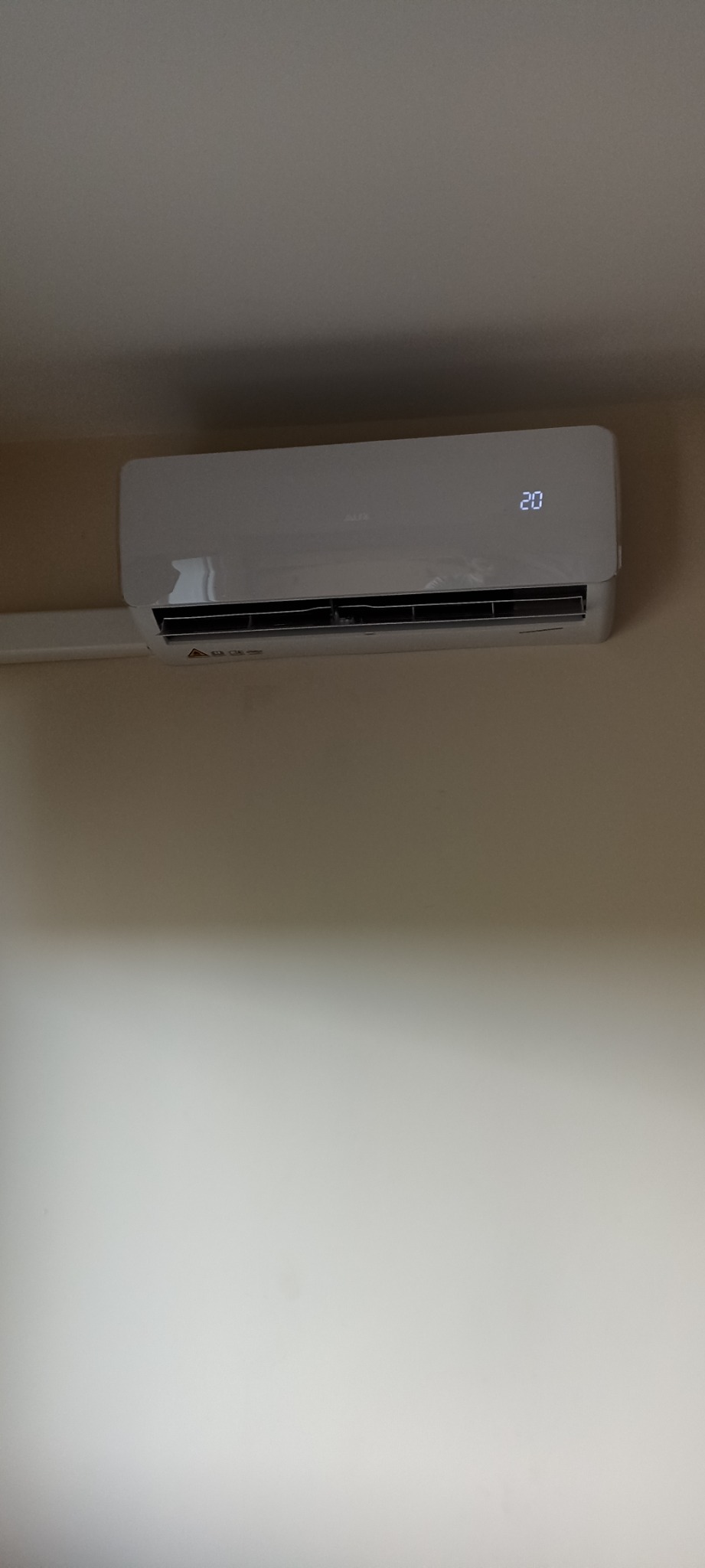 Biały klimatyzator ścienny marki AUX z wyświetlaczem temperatury 20 stopni zamontowany na beżowej ścianie.