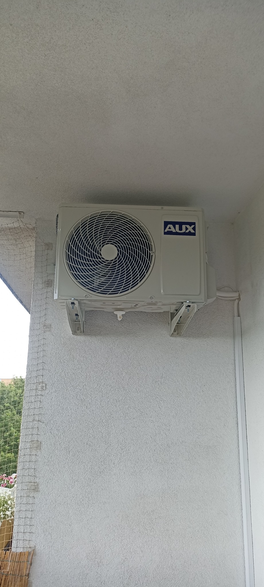 Biała jednostka zewnętrzna klimatyzatora marki AUX zamontowana na szarej, teksturowanej ścianie balkonu z siatką zabezpieczającą.