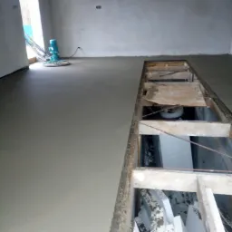 Świeżo wylana posadzka jastrychowa, częściowo jeszcze mokra, z widocznym otworem w podłodze zabezpieczonym drewnianą ramą i leżącymi wewnątrz płytami izolacyjnymi, obok stoi maszyna do zacierania...