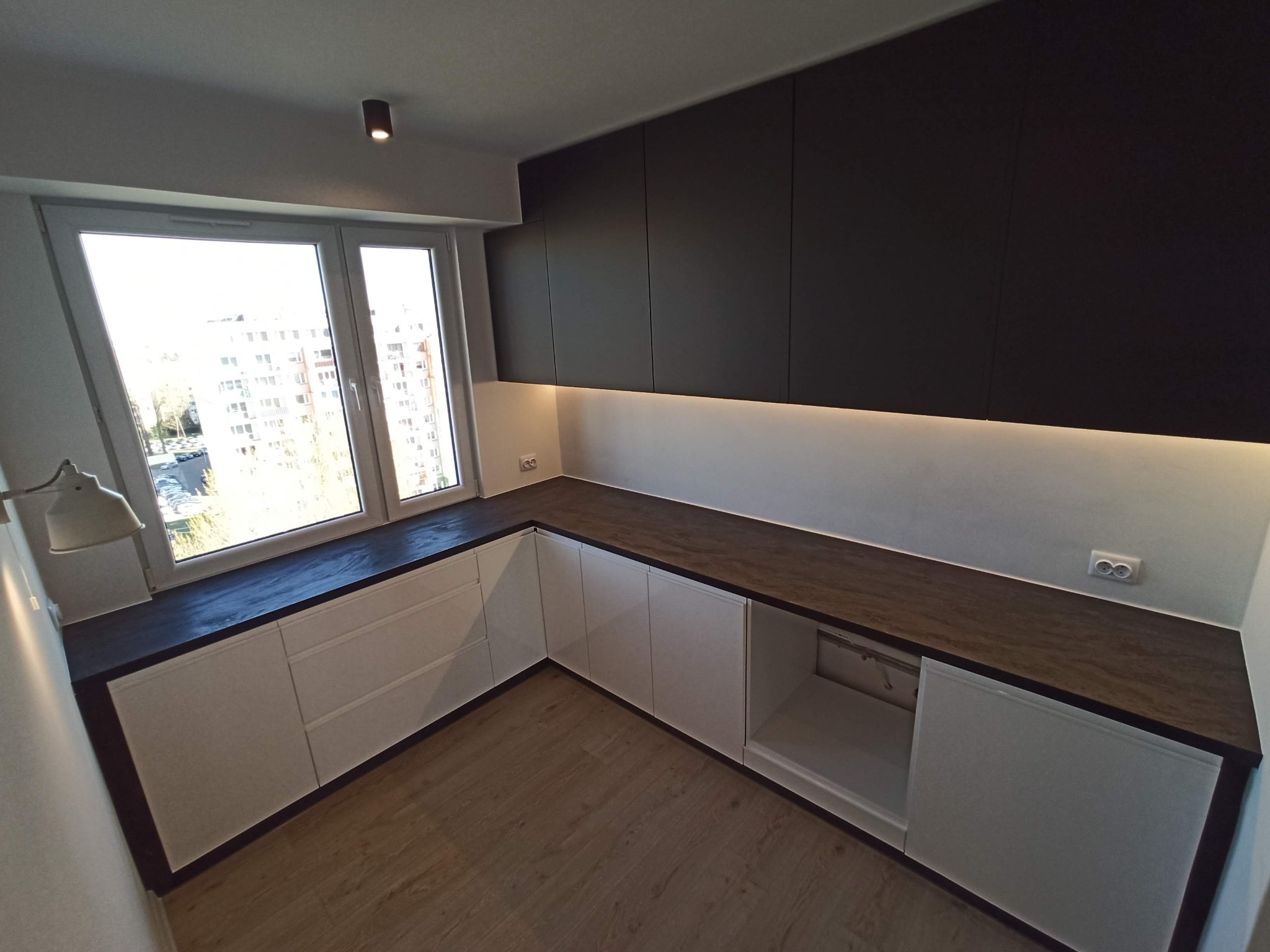 Nowoczesna kuchnia z białymi szafkami dolnymi i ciemnymi szafkami górnymi, oświetlenie LED pod szafkami, drewniana podłoga i widok z okna na budynki.