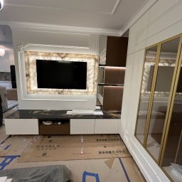 Unikkon Hanna Makowska - Nowoczesna zabudowa RTV z podświetlanym panelem z onyksu i szafkami w bieli. Na podłodze kartony zabezpieczające. Widoczna szafa z przeszklonymi drzwiami.