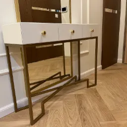 Biała konsola z trzema szufladami i złotymi uchwytami na tle parkietu w jodełkę, z widocznym fragmentem lustra i drzwi w tle. Nowoczesny design.