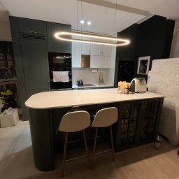 Unikkon Hanna Makowska - Nowoczesna kuchnia z wyspą, ciemnozielone szafki, biały blat z marmurowym wzorem, designerska lampa wisząca, dwie białe krzesła barowe, jasna podłoga.