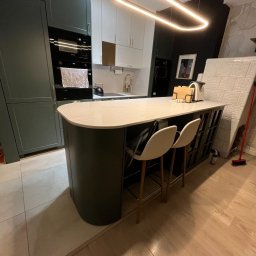 Unikkon Hanna Makowska - Nowoczesna kuchnia z wyspą: ciemnozielone szafki, biały blat, designerska lampa, krzesła barowe. Widok na zabudowę kuchenną z piekarnikiem i płytą.