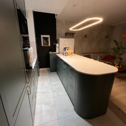 Unikkon Hanna Makowska - Nowoczesna kuchnia z wyspą, fronty w odcieniach zieleni, biały blat z marmurkowym wzorem. Oświetlenie ledowe nad wyspą. W tle salon.