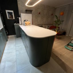 Unikkon Hanna Makowska - Nowoczesna wyspa kuchenna z zaokrąglonymi szafkami w kolorze grafitowym i blatem z białego kamienia. Nad nią designerska lampa. W tle salon z sofą i rośliną.