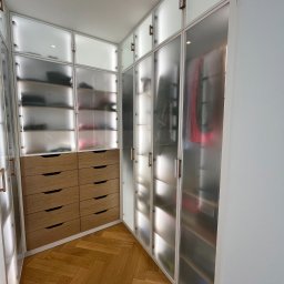 Unikkon Hanna Makowska - Funkcjonalna szafa wnękowa z drewnianymi szufladami i przesuwnymi, matowymi drzwiami, dopasowana do kąta pomieszczenia. Parkiet w jodełkę na podłodze.