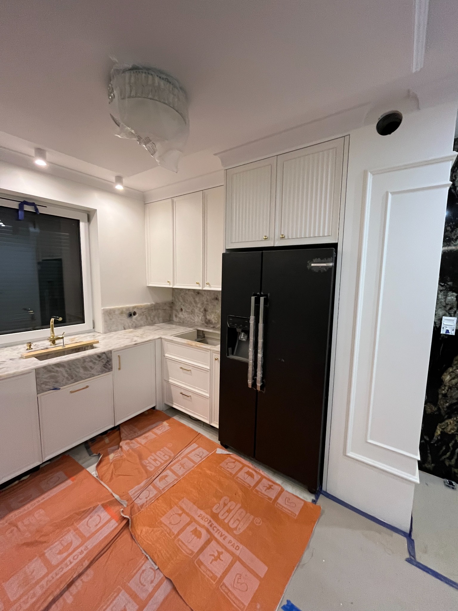 Nowoczesna, biała kuchnia z marmurowym blatem w trakcie montażu, zabezpieczona folią ochronną. Widoczna czarna lodówka i ozdobne listwy.