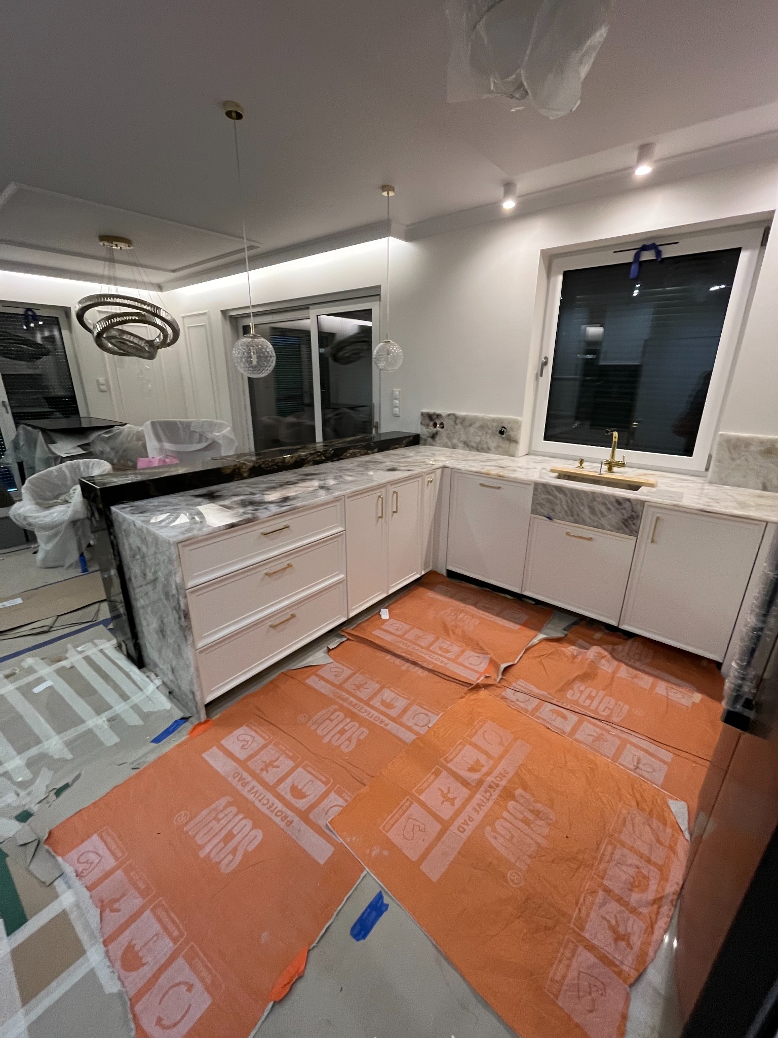 Jasna kuchnia z marmurowym blatem i złotą armaturą, zabezpieczona folią ochronną podczas montażu. Nowoczesne oświetlenie i okno w tle.