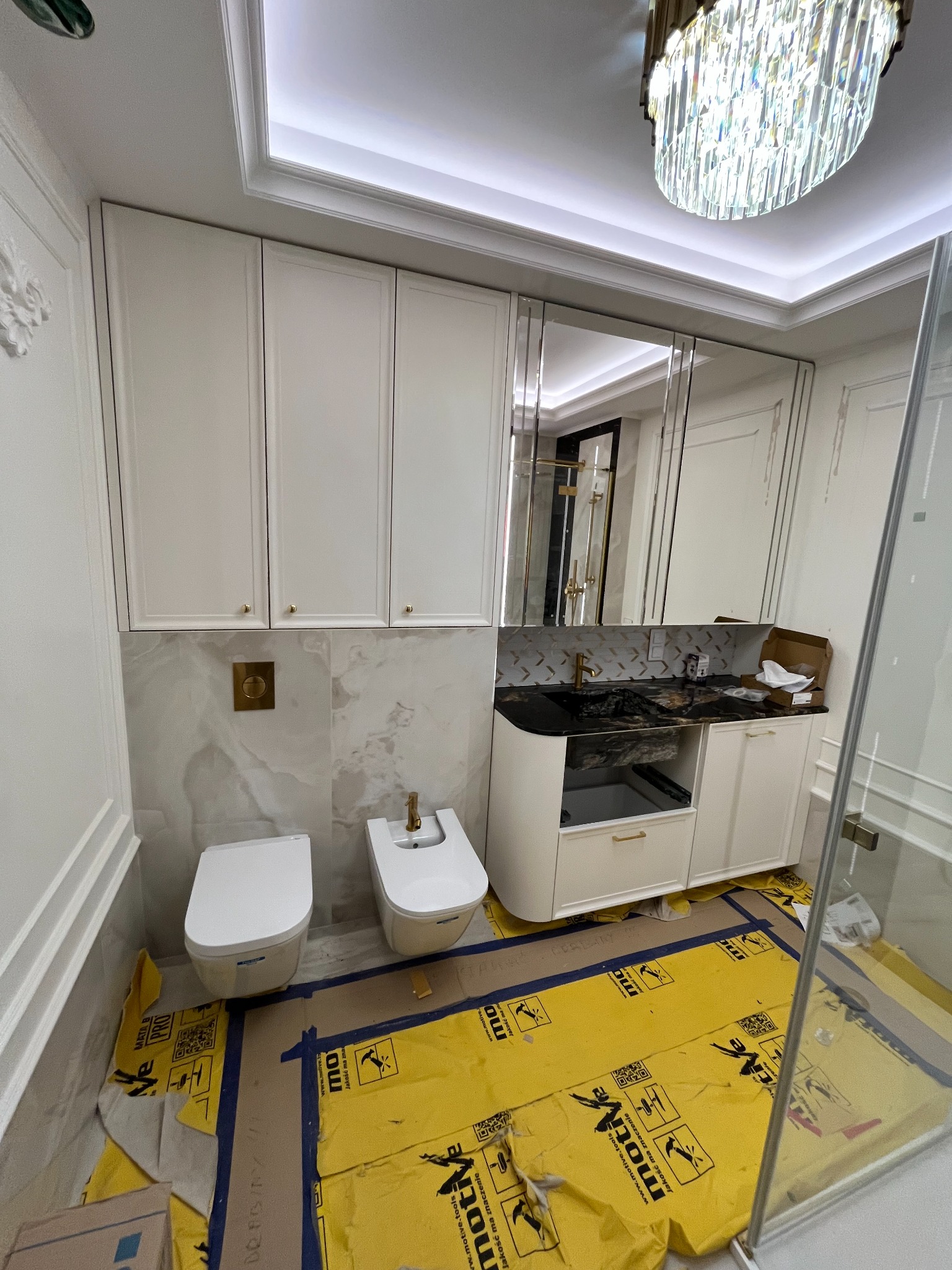 Elegancka łazienka w trakcie montażu: białe szafki wiszące i pod umywalką z czarnym blatem, toaleta i bidet, lustra w ramie. Oświetlenie sufitowe i zabezpieczona podłoga.