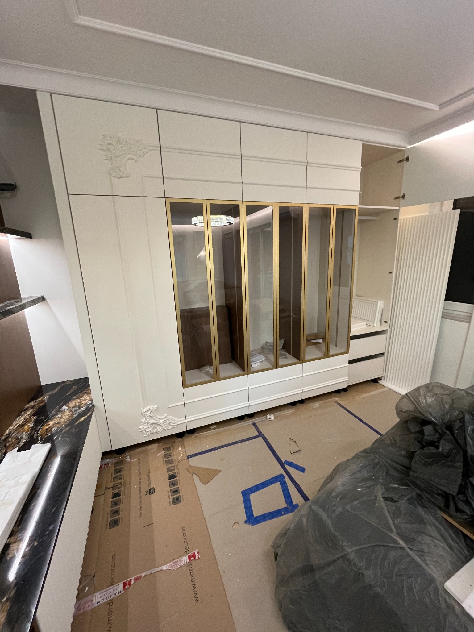 Biała szafa wnękowa z dekoracyjnymi detalami i złotymi ramkami szklanych drzwi, zamontowana w pomieszczeniu z zabezpieczoną podłogą.