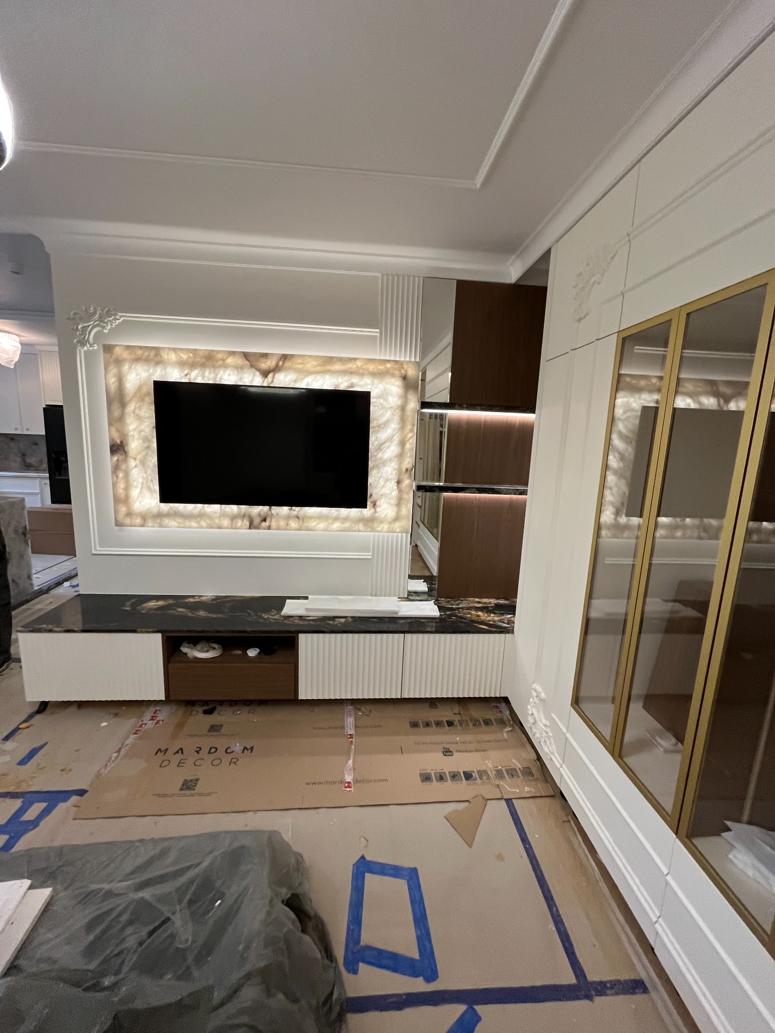 Nowoczesna zabudowa RTV z podświetlanym panelem z onyksu i szafkami w bieli. Na podłodze kartony zabezpieczające. Widoczna szafa z przeszklonymi drzwiami.