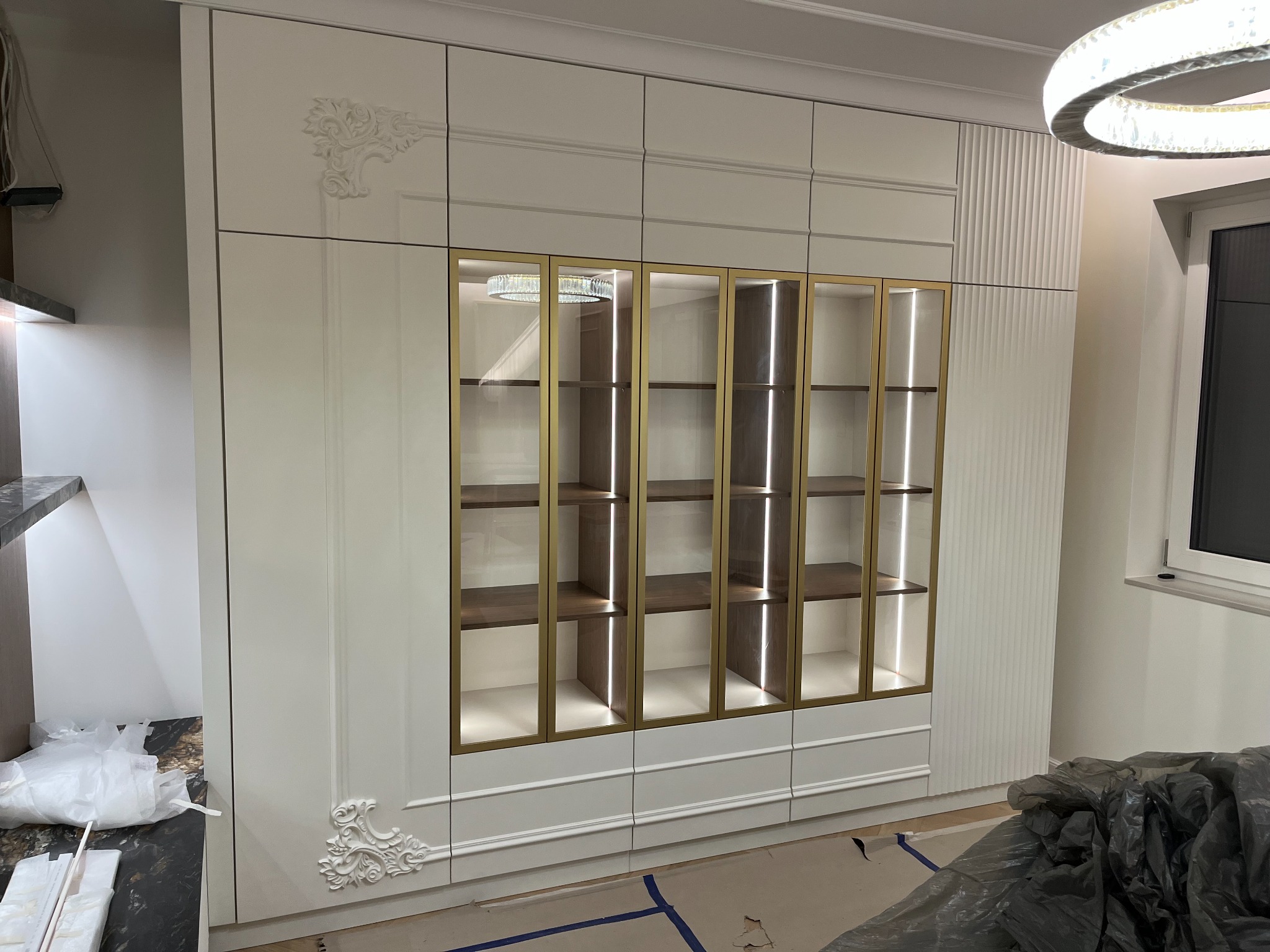 Biała, elegancka szafa wnękowa z przeszklonymi witrynami i złotymi ramkami, zdobiona ornamentami. Widoczne półki i oświetlenie LED. Część pomieszczenia w trakcie prac wykończeniowych.
