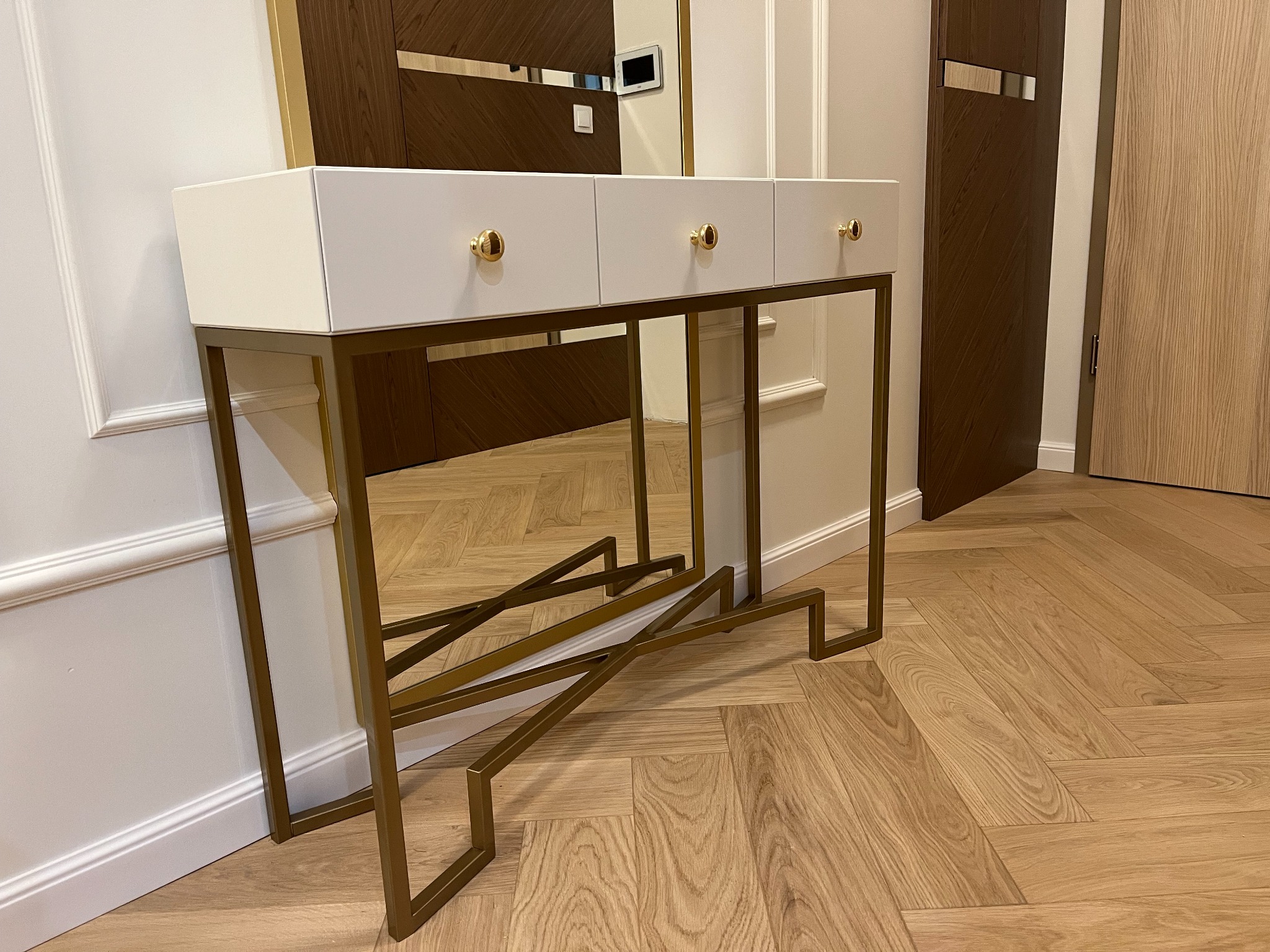 Biała konsola z trzema szufladami i złotymi uchwytami na tle parkietu w jodełkę, z widocznym fragmentem lustra i drzwi w tle. Nowoczesny design.