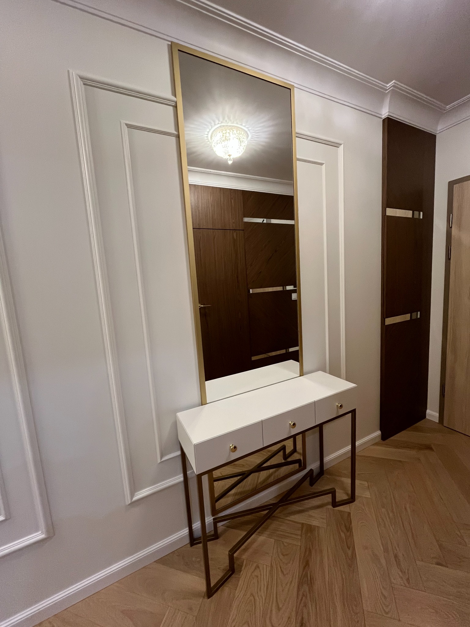 Biała konsola z trzema szufladami i złotymi uchwytami, zwieńczona lustrem w złotej ramie, na tle ściany z dekoracyjnymi listwami i parkietu w jodełkę.