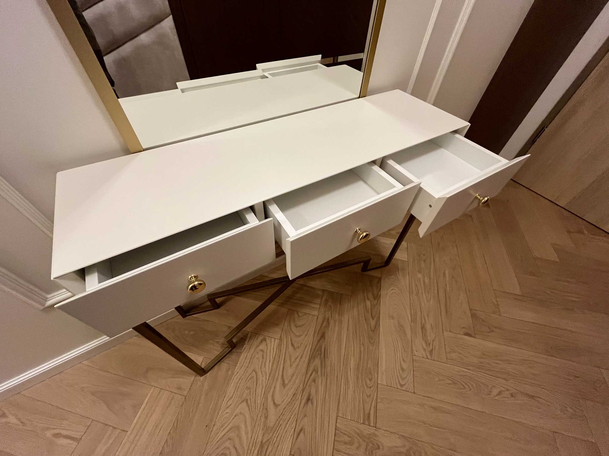Biała toaletka z lustrem w złotej ramie, otwarte szuflady z okrągłymi, złotymi uchwytami, parkiet w jodełkę, nowoczesny design, perspektywa z góry.