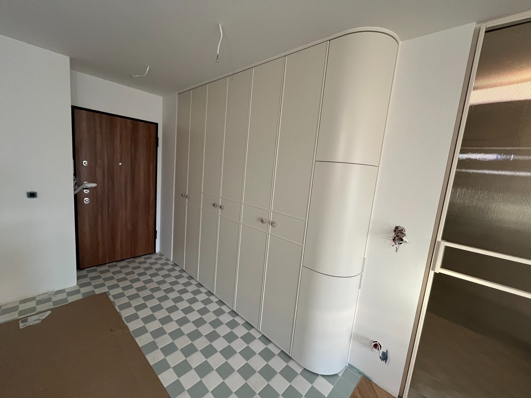 Biała szafa wnękowa z zaokrąglonym bokiem, dopasowana do wnętrza z szachownicową podłogą i brązowymi drzwiami wejściowymi. Widoczne niedokończone detale.