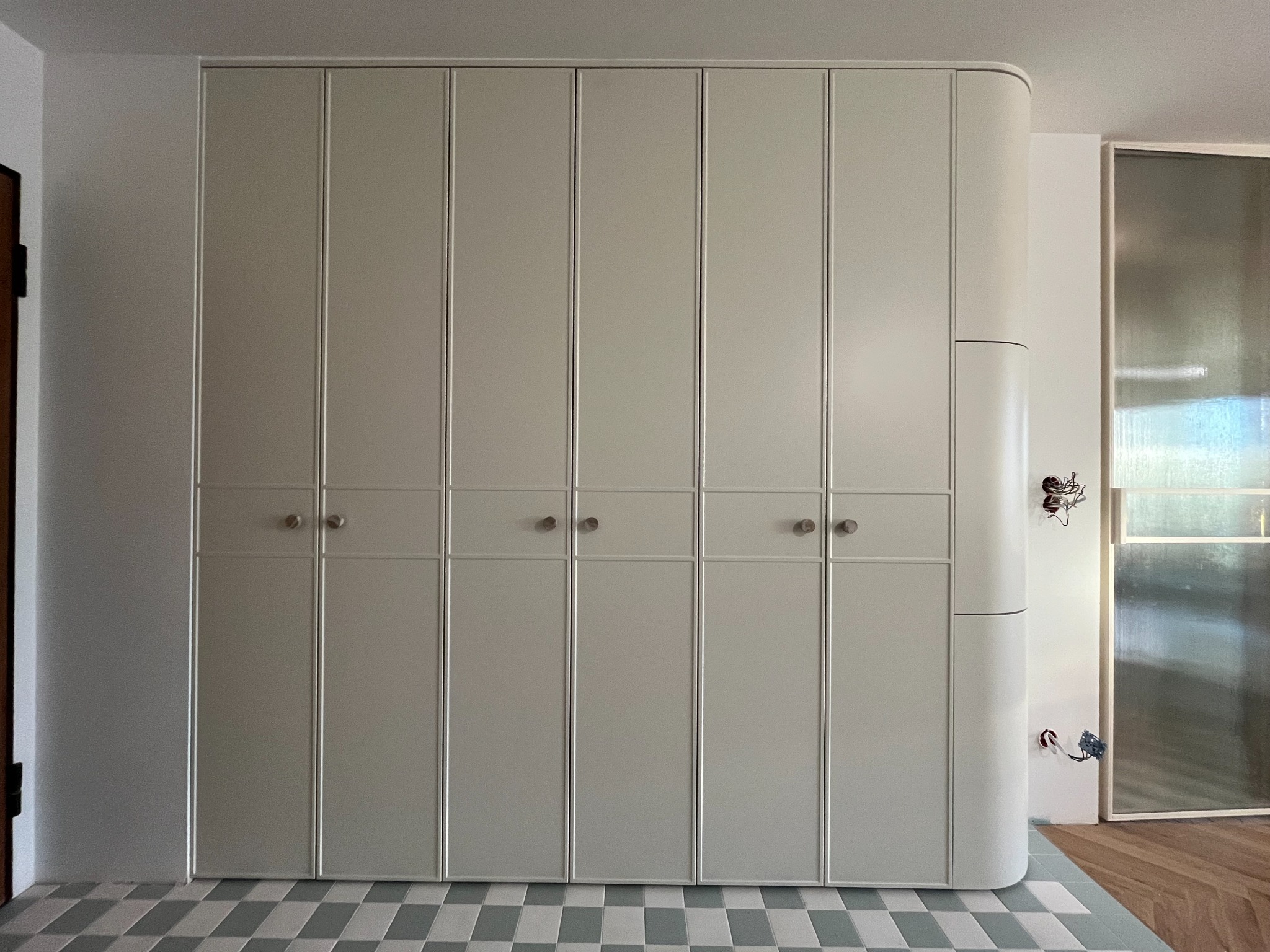 Jasnoszara szafa wnękowa z zaokrąglonym bokiem, widoczna na tle szachownicy podłogowej i fragmentu drzwi.