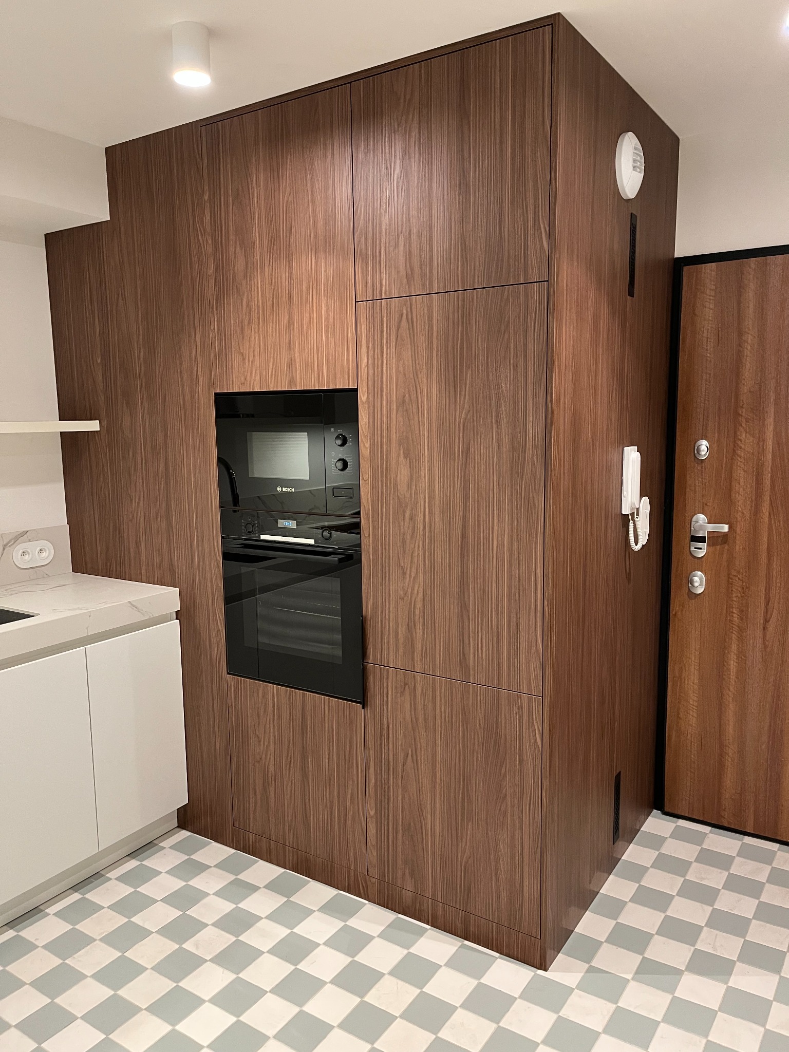 Nowoczesna zabudowa kuchenna z forniru orzechowego, z wbudowanym piekarnikiem Bosch i białym blatem. Podłoga w szachownicę, drzwi wejściowe z domofonem.