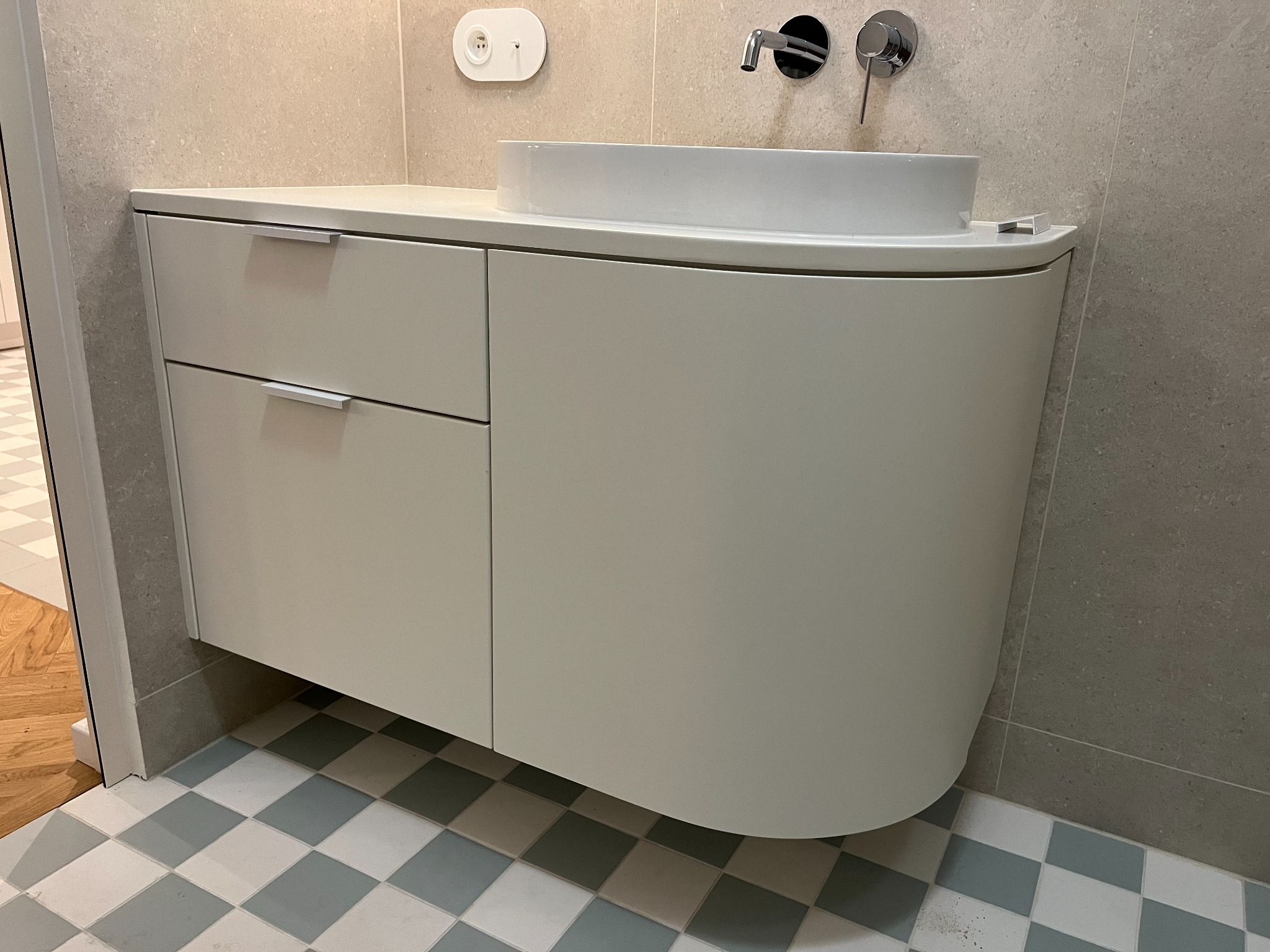 Biała szafka łazienkowa z umywalką nablatową i chromowaną armaturą na tle szarej ściany i podłogi w biało-szarą szachownicę.