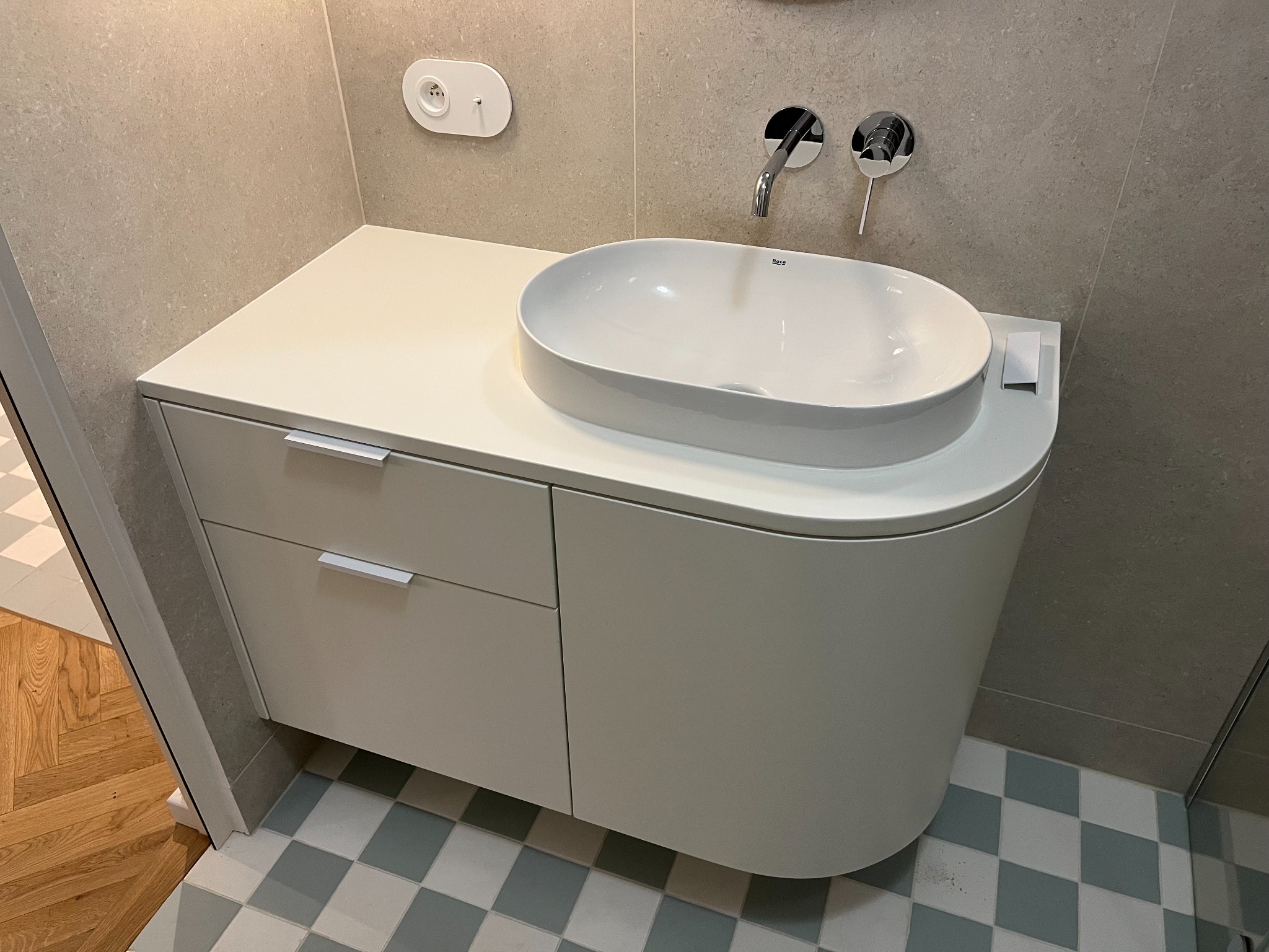 Biała szafka łazienkowa z owalną umywalką Roca, chromowana bateria ścienna, szaro-beżowe płytki i podłoga w szachownicę z akcentem niebieskiego.