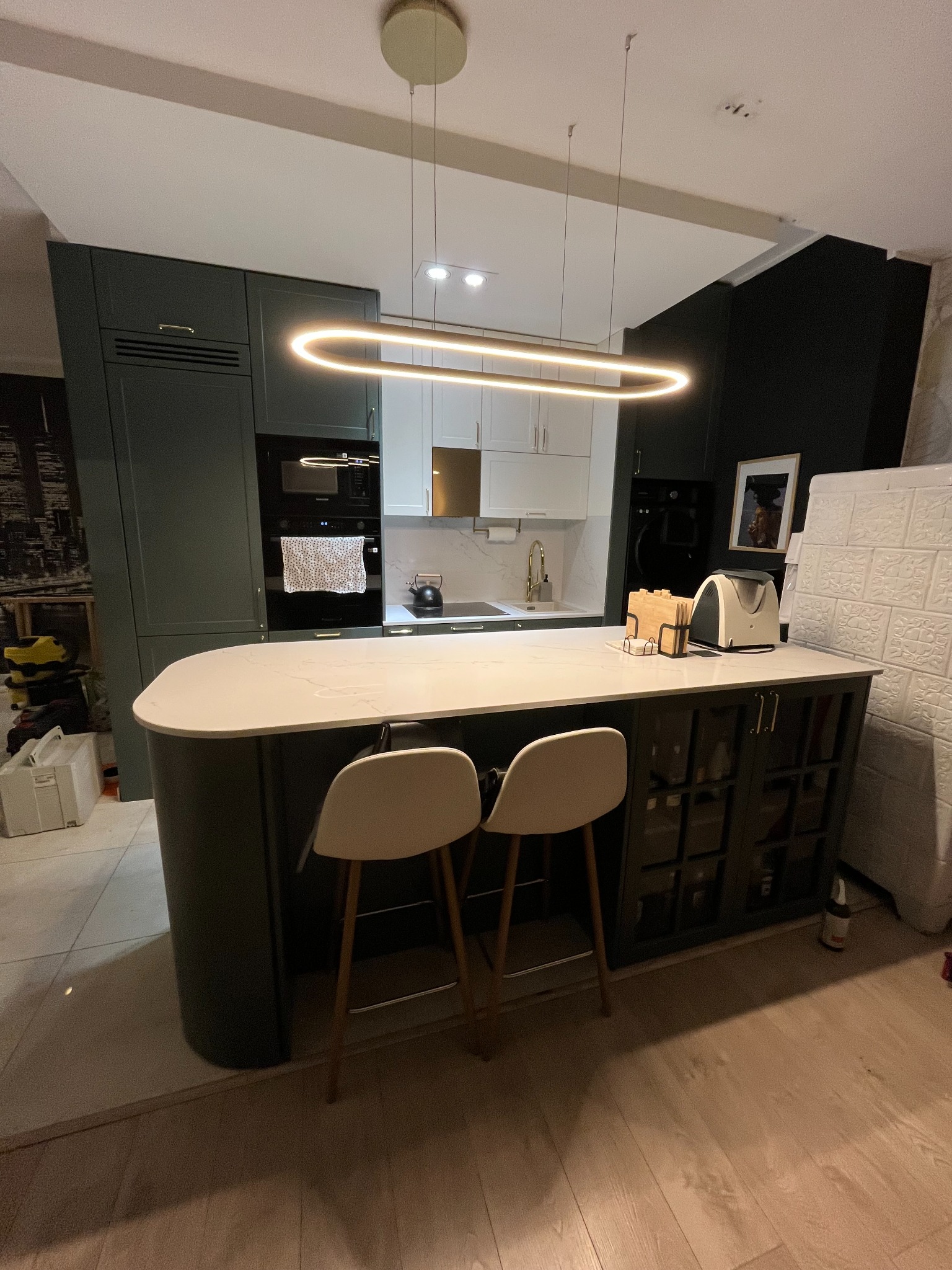 Nowoczesna kuchnia z wyspą, ciemnozielone szafki, biały blat z marmurowym wzorem, designerska lampa wisząca, dwie białe krzesła barowe, jasna podłoga.