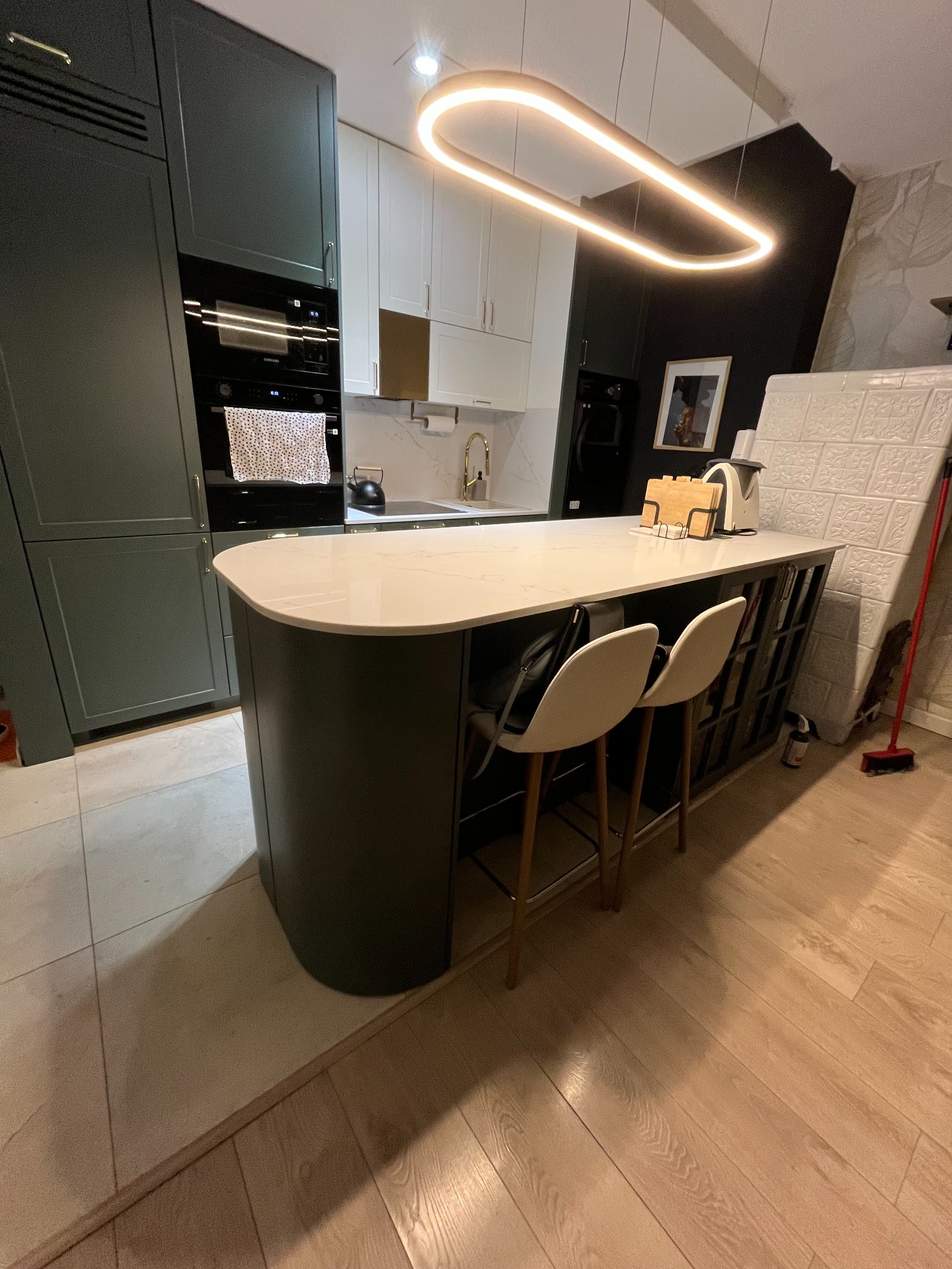 Nowoczesna kuchnia z wyspą: ciemnozielone szafki, biały blat, designerska lampa, krzesła barowe. Widok na zabudowę kuchenną z piekarnikiem i płytą.