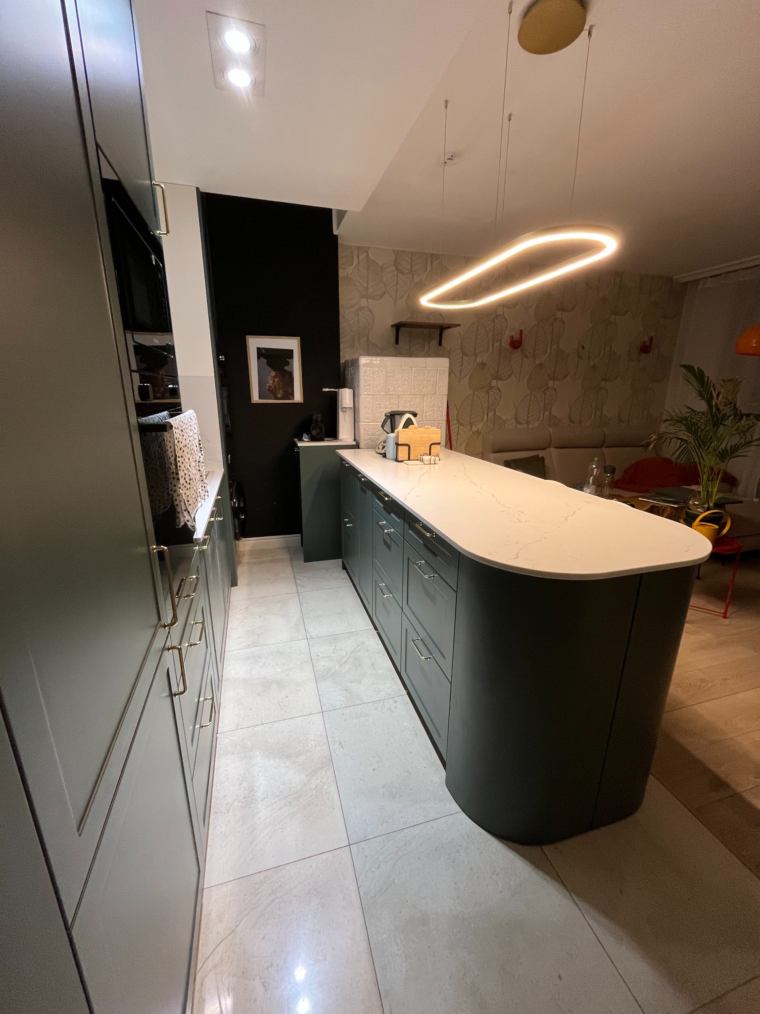 Nowoczesna kuchnia z wyspą, fronty w odcieniach zieleni, biały blat z marmurkowym wzorem. Oświetlenie ledowe nad wyspą. W tle salon.