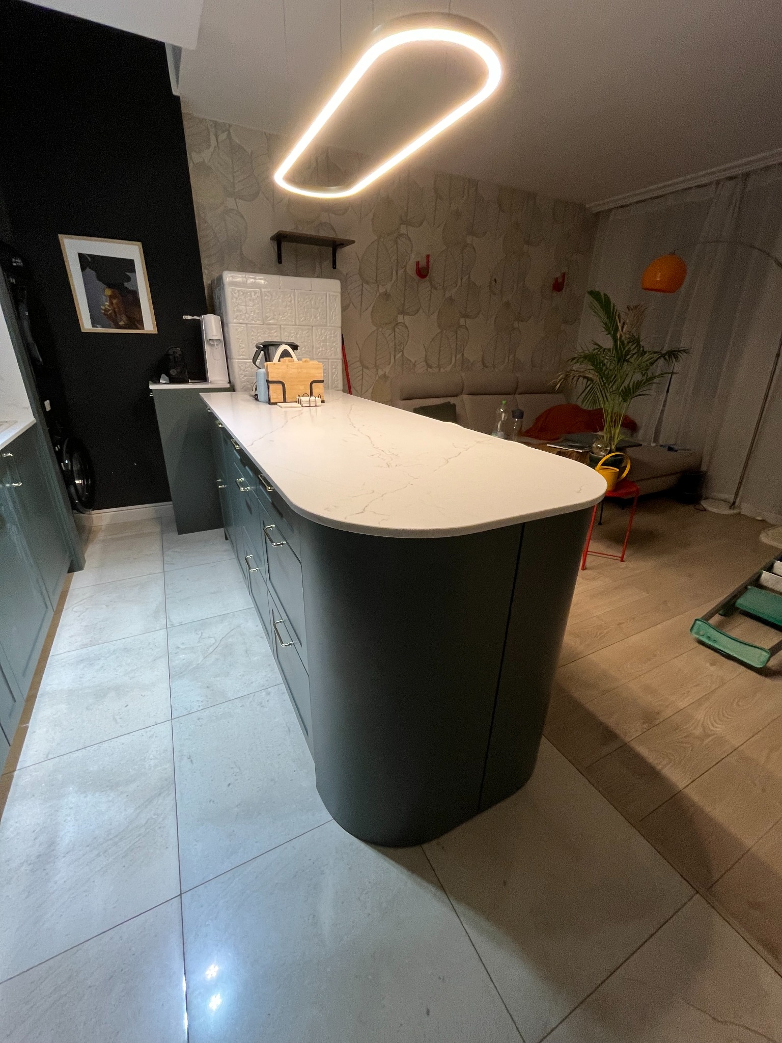 Nowoczesna wyspa kuchenna z zaokrąglonymi szafkami w kolorze grafitowym i blatem z białego kamienia. Nad nią designerska lampa. W tle salon z sofą i rośliną.