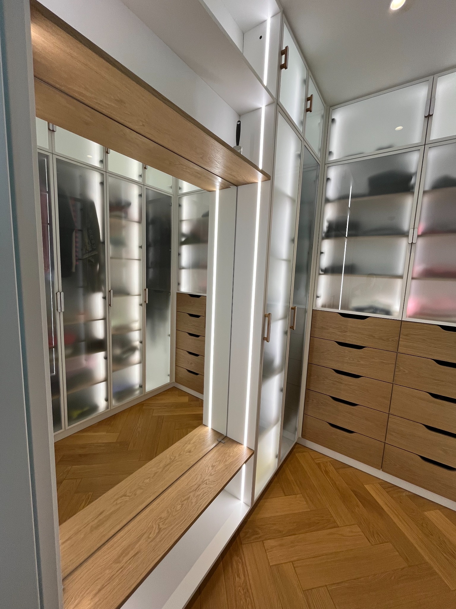 Nowoczesna garderoba z podświetlanymi półkami i szufladami z jasnego drewna, widok z perspektywy wejścia, parkiet w jodełkę na podłodze.