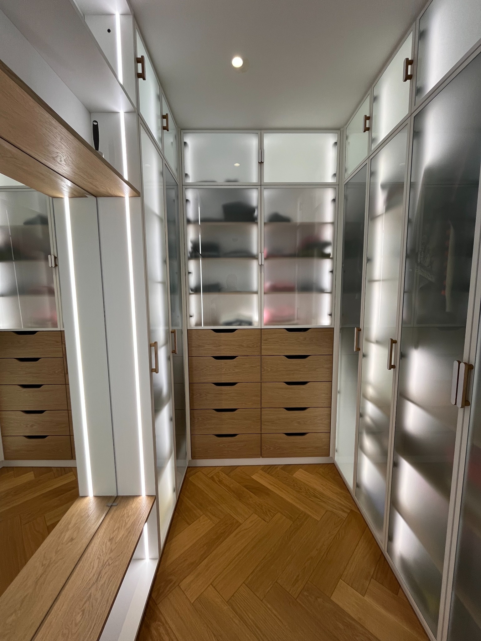 Nowoczesna garderoba z drewnianymi szufladami i matowymi, podświetlanymi drzwiami, parkiet w jodełkę. Minimalistyczny design, jasne wnętrze, eleganckie wykończenie.