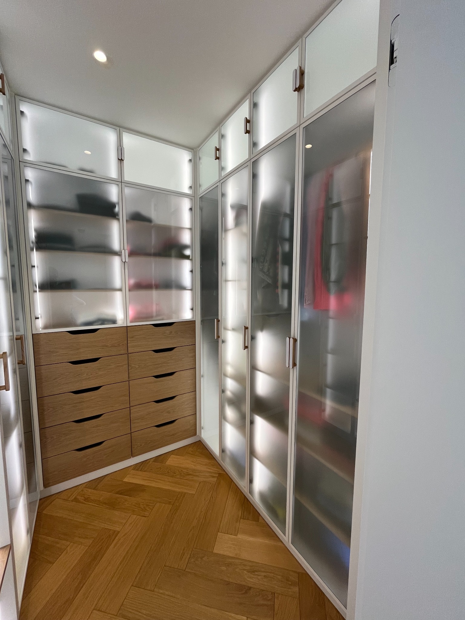 Funkcjonalna szafa wnękowa z drewnianymi szufladami i przesuwnymi, matowymi drzwiami, dopasowana do kąta pomieszczenia. Parkiet w jodełkę na podłodze.