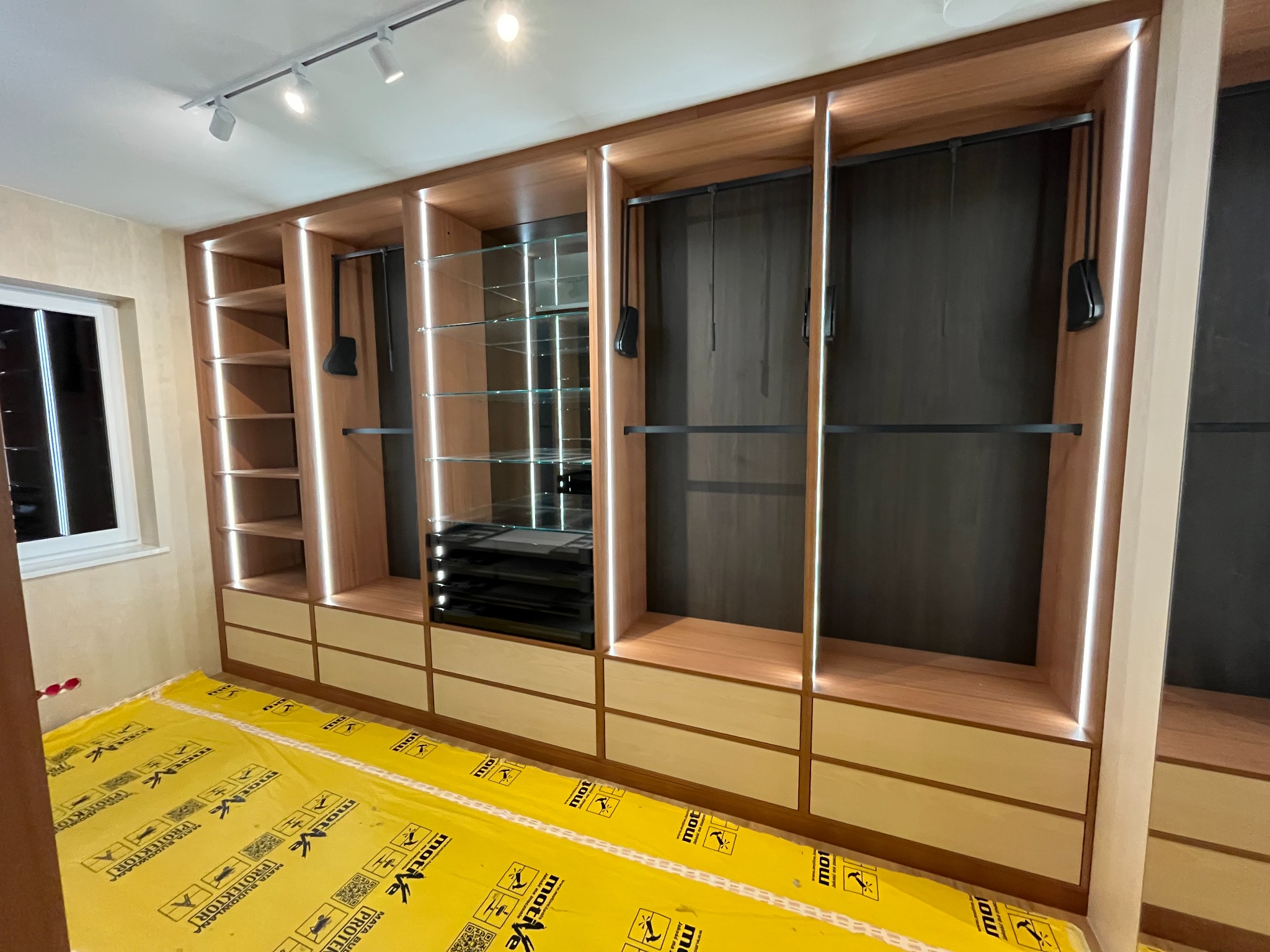 Nowoczesna szafa wnękowa z oświetleniem LED i szklanymi półkami, zabezpieczona folią ochronną na podłodze, widok z dołu.