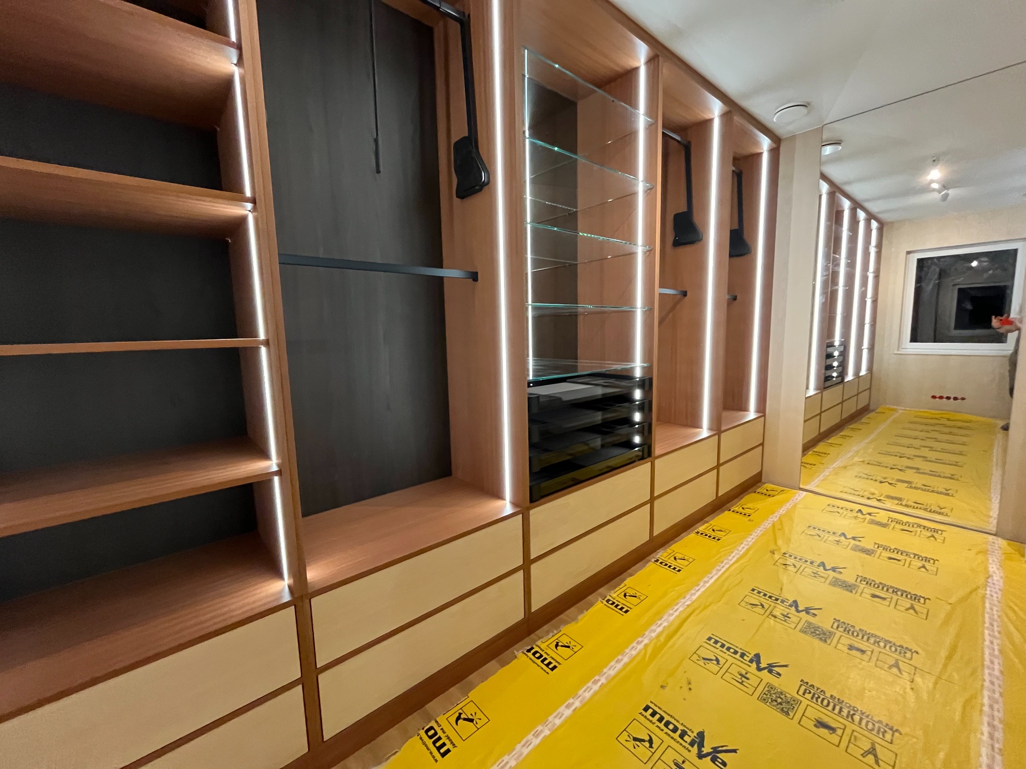 Nowoczesna garderoba z oświetleniem LED, drewnianymi półkami i szufladami w odcieniach beżu i brązu. Widoczne elementy wyposażenia i zabezpieczenie podłogi na czas montażu.