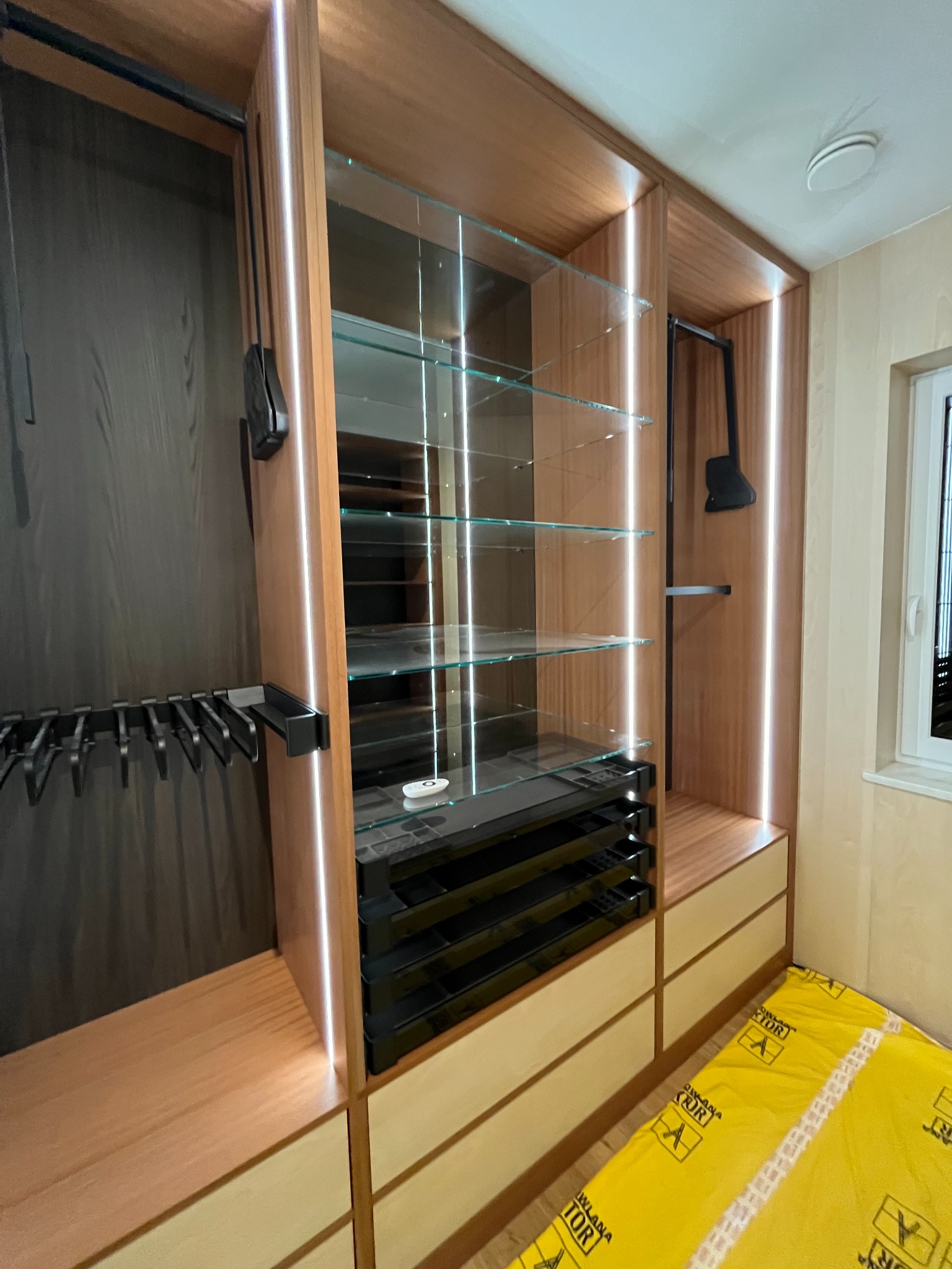 Nowoczesna szafa wnękowa z oświetleniem LED, szklanymi półkami i wysuwanymi szufladami. Widoczne detale wykończenia i akcesoria do przechowywania ubrań.