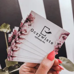 Stos wizytówek z kwiatowym motywem i logo firmy 'Oczywiste stylizacja oka' trzymany w dłoni z koralowymi paznokciami na tle czarno-białych pasów i rośliny.