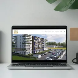 Laptop wyświetla stronę internetową z wizualizacją nowoczesnego budynku mieszkalnego z parkingiem, obok ramka na zdjęcia i aparat retro.