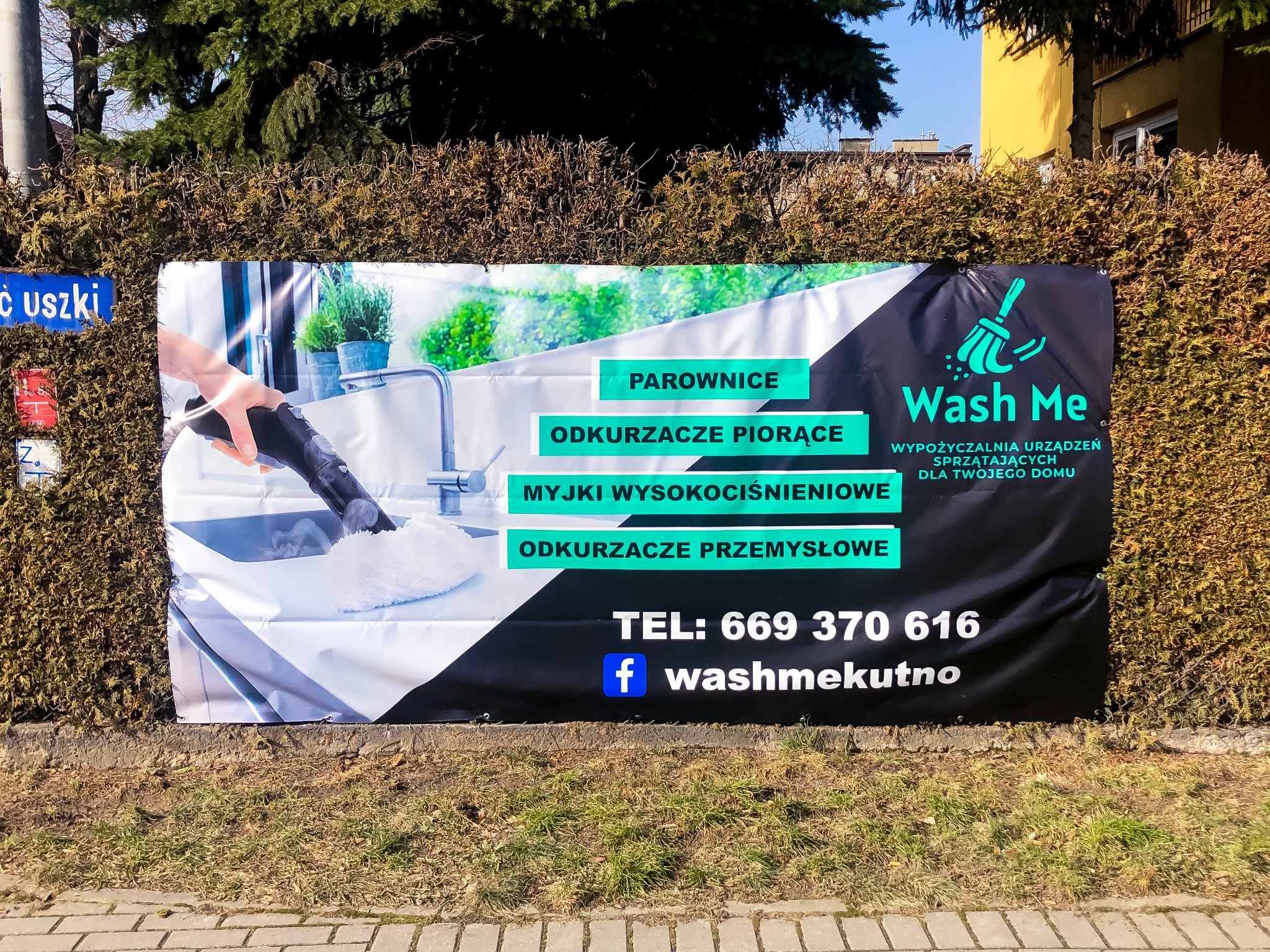 Baner reklamowy firmy Wash Me z ofertą wynajmu urządzeń sprzątających, przedstawiający mycie zlewu parownicą, umieszczony na żywopłocie przy ulicy.