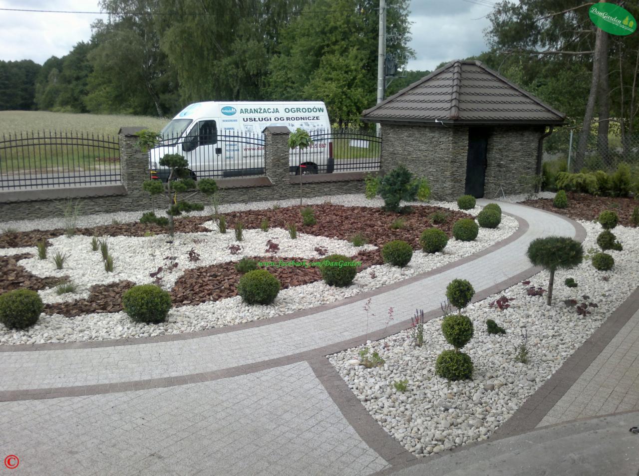 Aranżacja ogrodu z ścieżką z kostki, białym żwirem i ozdobnymi krzewami. W tle ogrodzenie i budynek gospodarczy. Usługi ogrodnicze.