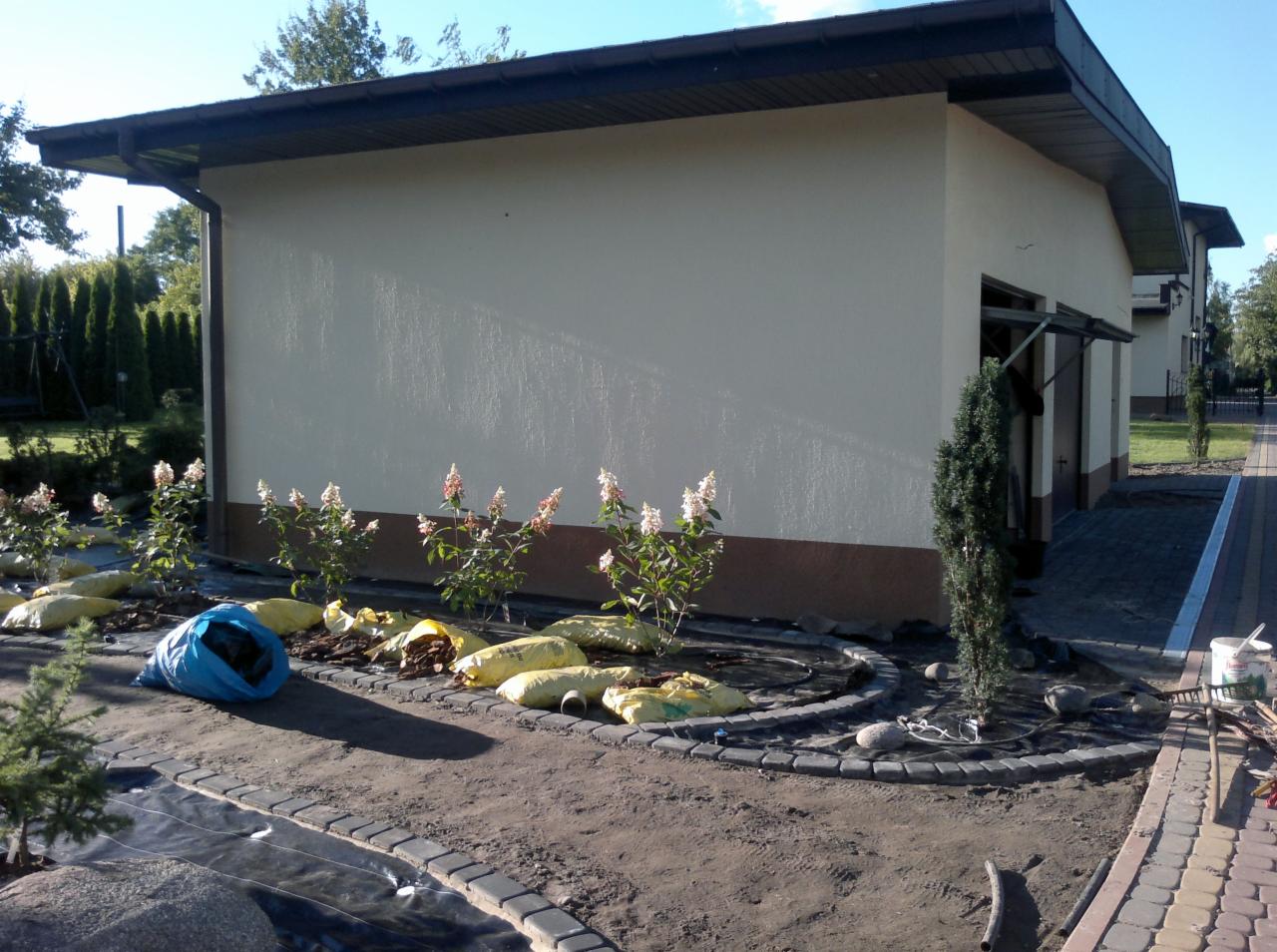 Ogród w trakcie aranżacji: nasadzenia hortensji bukietowych, obrzeża z kostki, worki z korą. Widoczny budynek w tle i narzędzia ogrodnicze.