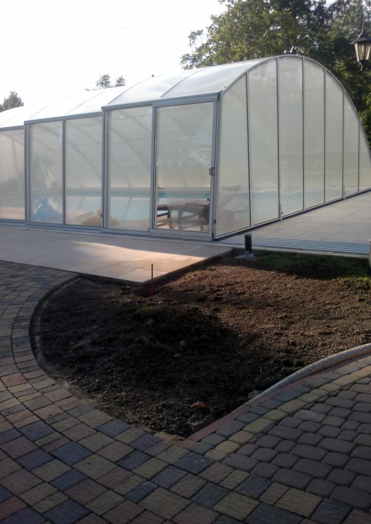 Zadaszenie basenu z przesuwnymi drzwiami, widoczne z poziomu brukowanej ścieżki i przygotowanego podłoża pod trawnik. Lekkie pochylenie kamery.