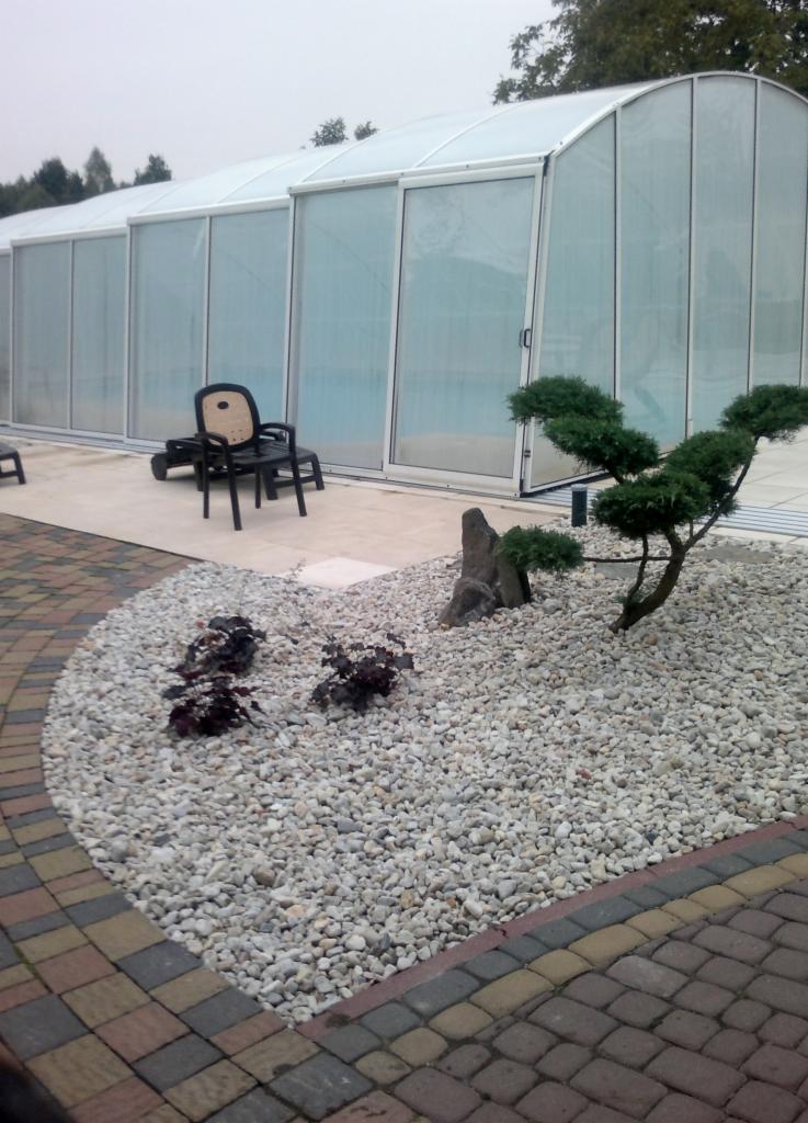 Ogród z białymi kamieniami i bonsai, obok zadaszony basen. Na tarasie krzesło ogrodowe. Kostka brukowa w tle. Specyficzny układ kamieni i roślin.