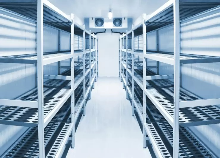 Pusta mroźnia z rzędami metalowych regałów z perforowanymi półkami i klimatyzatorem sufitowym z dwoma wentylatorami, oświetlona chłodnym, jasnym światłem.