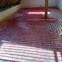 Czerwone rury systemu grzewczego ułożone spiralnie na srebrnej folii izolacyjnej, zamocowane do metalowej siatki w trakcie instalacji ogrzewania podłogowego na poddaszu.