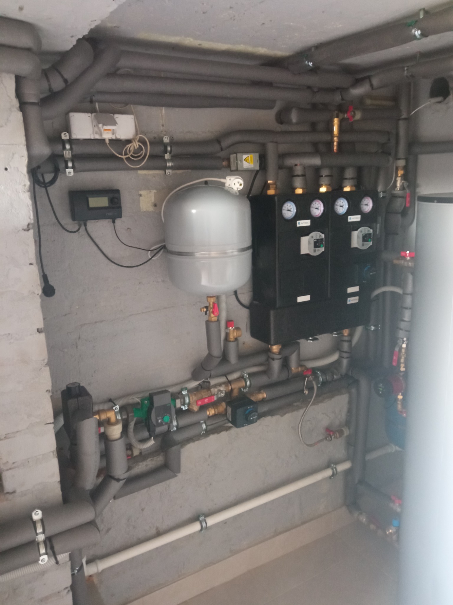 Rozbudowana instalacja grzewcza w piwnicy z widocznymi rurami izolowanymi termicznie, manometrami, zbiornikiem wyrównawczym i sterownikiem elektronicznym na szarej, surowej ścianie.