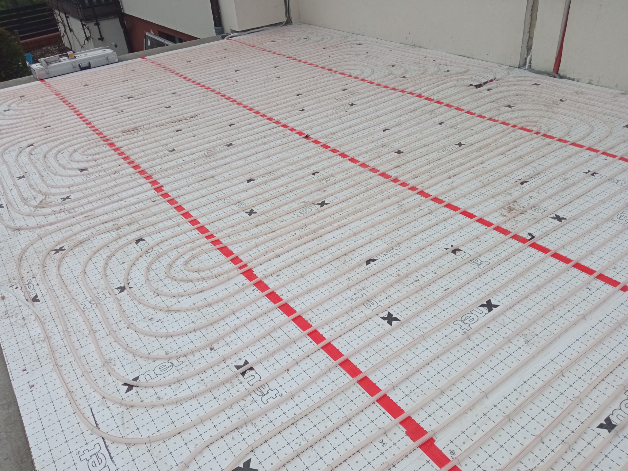 Rozłożona instalacja ogrzewania podłogowego Xnet na białej macie izolacyjnej z czerwonymi prowadnicami, widok z góry, przygotowana do zalania wylewką.