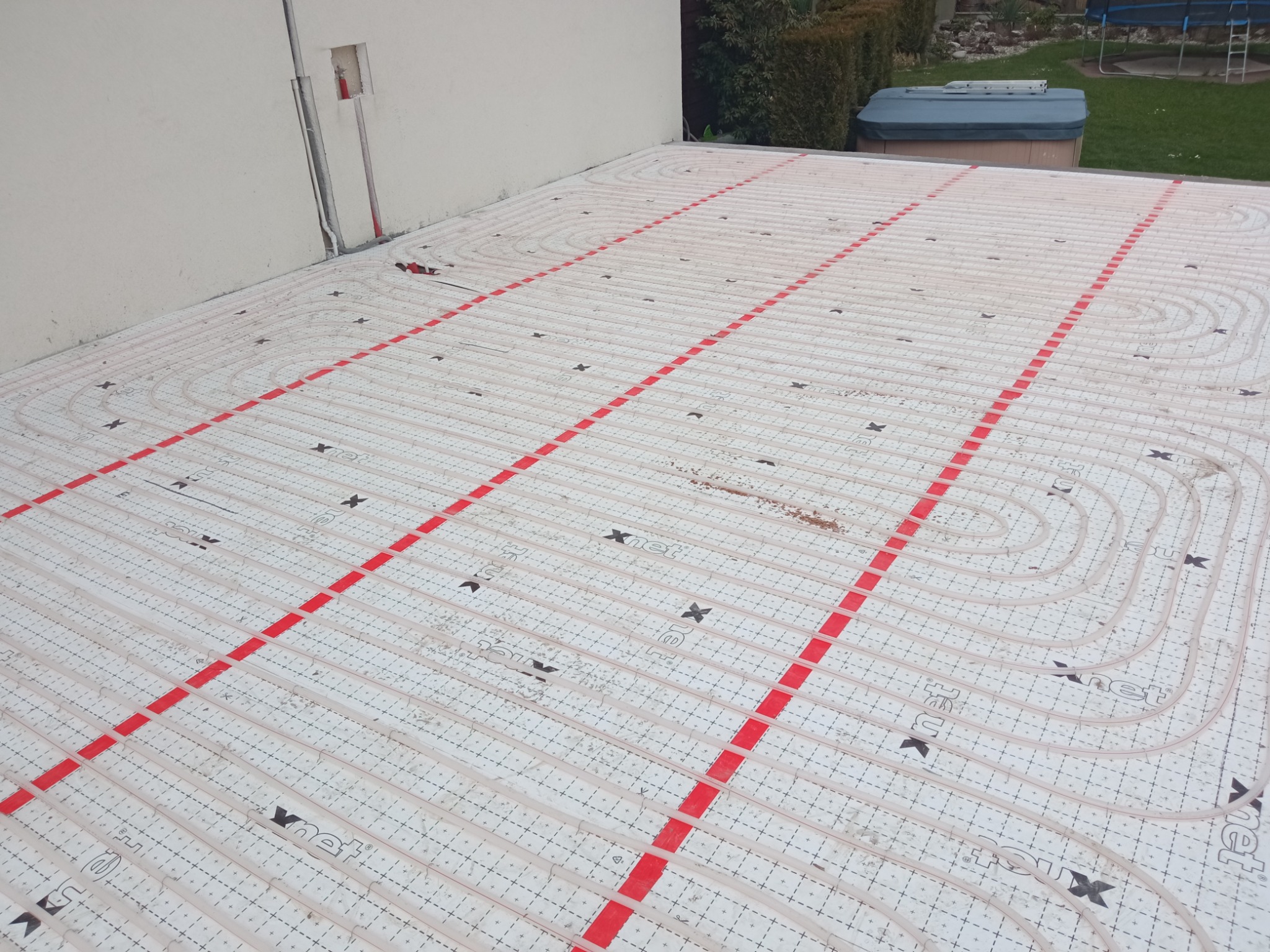 Rozłożona instalacja ogrzewania podłogowego Xnet na białej macie z czerwonymi liniami, widoczne rury i elementy montażowe, w tle ściana budynku i fragment ogrodu z jacuzzi.